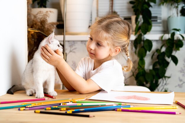 Una bambina bionda si siede al tavolo e abbraccia dolcemente un gattino scozzese bianco e sorride colorato