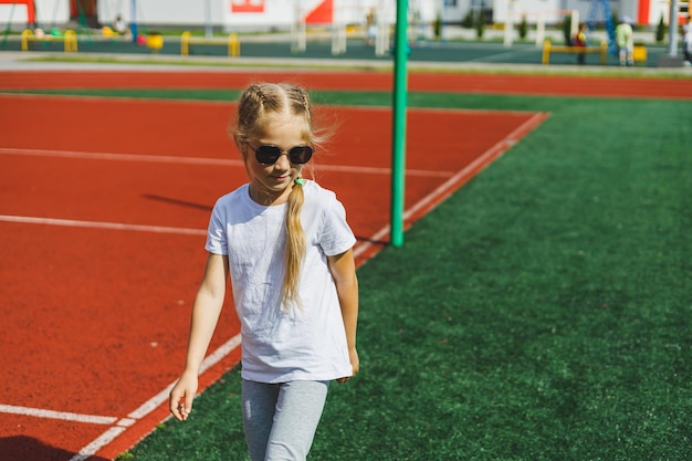 Una bambina bionda sta correndo sul campo sportivo e sorridente indossa occhiali da sole e una maglietta bianca Vacanze estive e tempo di vacanza