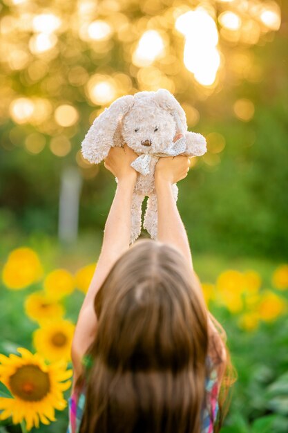 Una bambina cammina in un campo di girasoli e tiene in mano un coniglio di peluche