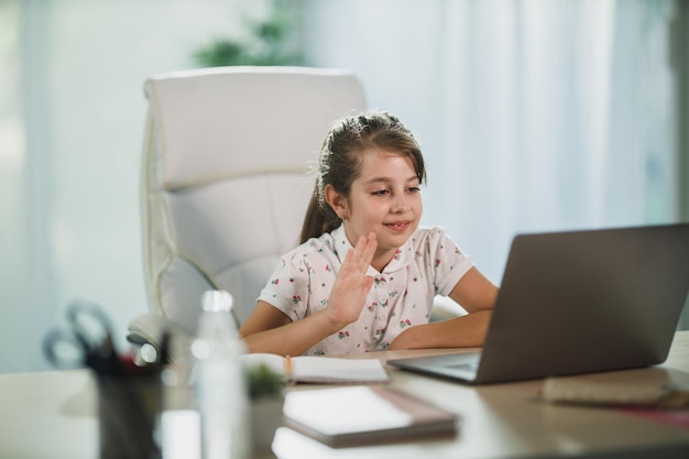 Una bambina carina che usa il suo laptop per fare una lezione online e saluta l'insegnante durante una videochiamata.