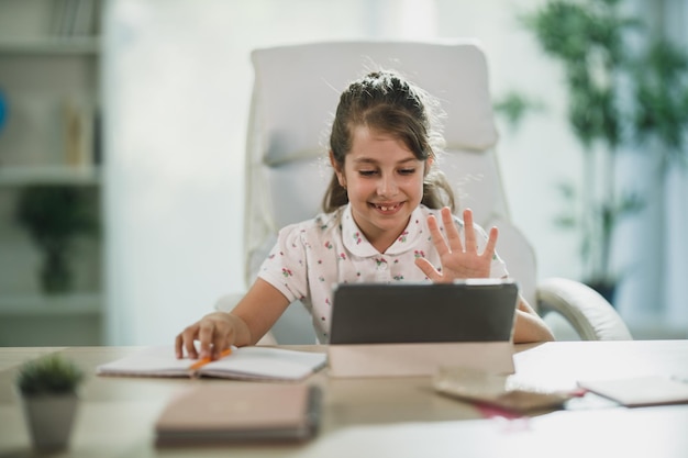 Una bambina carina che usa il suo tablet digitale per fare una lezione online e saluta l'insegnante durante una videochiamata.