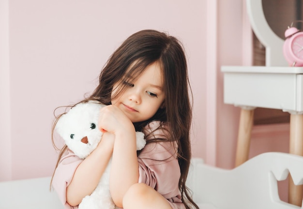 Una bambina carina con i capelli lunghi abbraccia un orsacchiotto guarda la telecamera e sorride mentre è seduta sul suo letto a casa