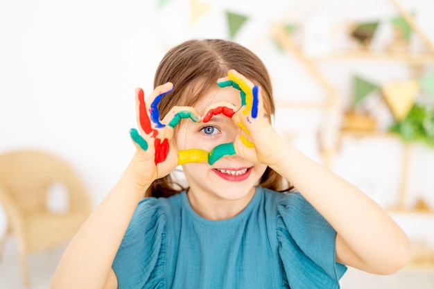 una bambina carina con le mani colorate dipinte con le dita mostra un cuore nella nursery e impara a disegnare o a giocare con le lezioni di arte con matite e vernici in primo piano