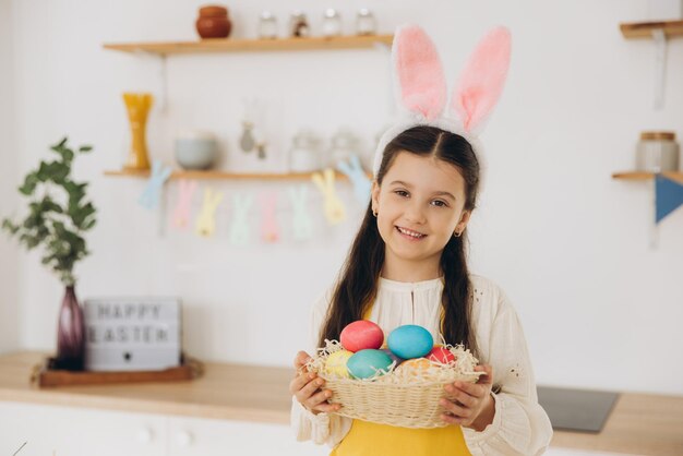 Una bambina carina con le orecchie di coniglietto di peluche e un grembiule che tiene un cesto pieno di uova colorate di Pasqua.