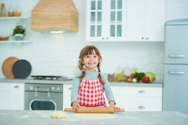 Una bambina carina con le trecce in un grembiule scozzese impasta l'impasto della pizza da sola. Corsi di cucina per bambini.