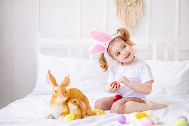 Una bambina carina con orecchie da coniglio e uova colorate su un letto bianco a casa giocando e sorridendo un bambino biondo celebra la buona Pasqua