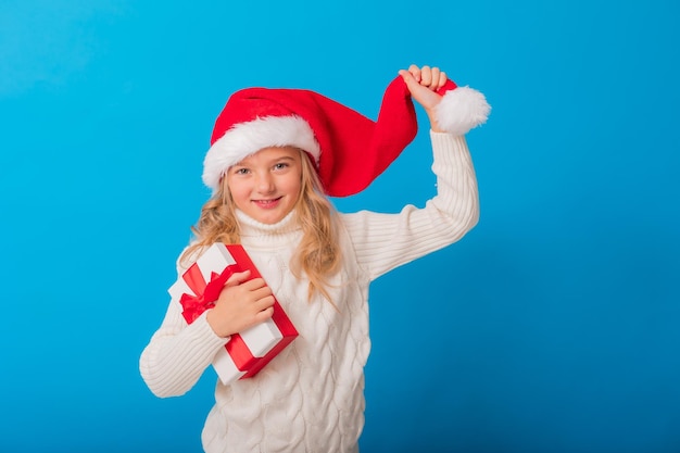 Una bambina carina con un cappello da Babbo Natale tiene un regalo su uno sfondo blu nello studio Spazio per il testo Maglione lavorato a maglia concetto invernale