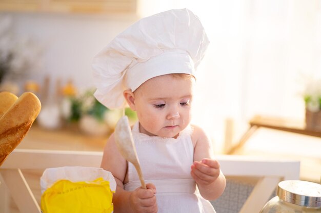 Una bambina carina con un cappello da chef e un grembiule è seduta sul tavolo in cucina a casa con un mattarello farina pagnotte di pane un piccolo cuoco cibo fatto in casa