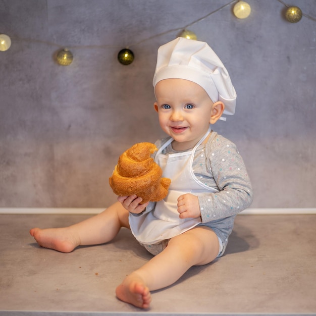 Una bambina carina con un cappello da chef e un grembiule si siede a casa sul tavolo in cucina