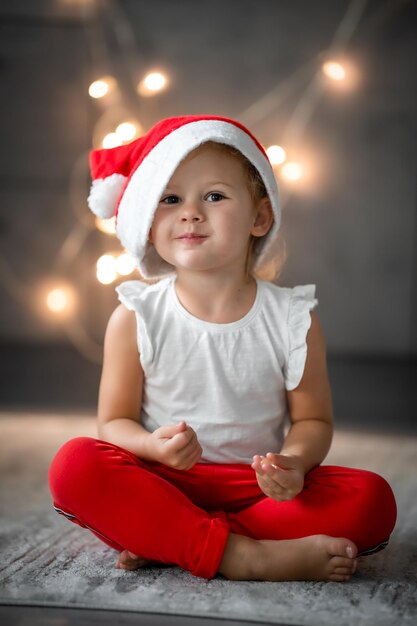 Una bambina carina con un cappello rosso e una camicia bianca Concetto natalizio con bambino e ghirlanda sullo sfondo in sfocatura