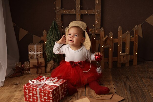 una bambina carina con un costume di Capodanno e un cappello con paraorecchie è seduta accanto a un regalo