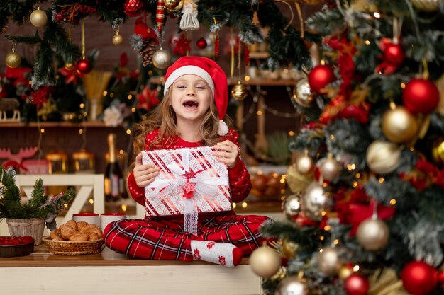 Una bambina carina con un maglione rosso e un cappello di Babbo Natale in una cucina buia con un albero di Natale si rallegra con i regali e aspetta il nuovo anno o il Natale