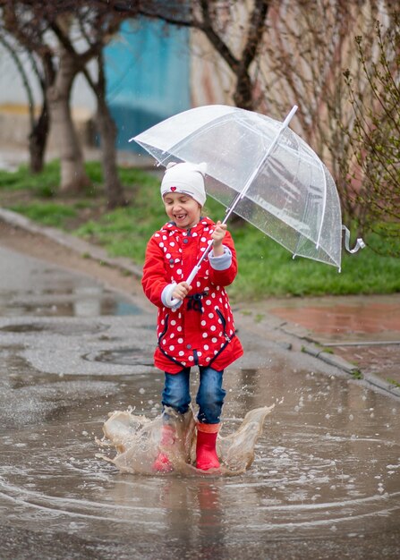 Una bambina carina con un mantello rosso stivali rossi e un cappello bianco salta nelle pozzanghere e si diverte