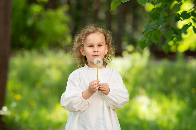 Una bambina carina con una camicia bianca tiene in mano un fiore di tarassaco e ci soffia sopra