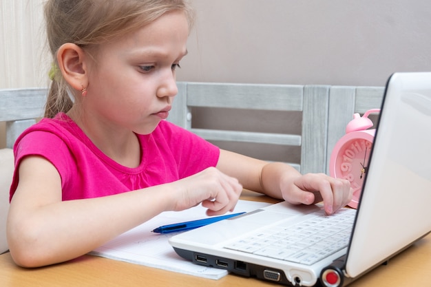 Una bambina carina è seduta a un tavolo e scrive i compiti su un laptop da inviare all'insegnante per la revisione. Concetto di apprendimento, apprendimento a distanza, autoeducazione, lezione di videoconferenza con mentore