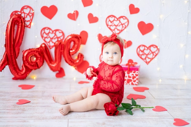 Una bambina carina è seduta in un body rosso su uno sfondo di cuori rossi e l'iscrizione ama il concetto di san valentino