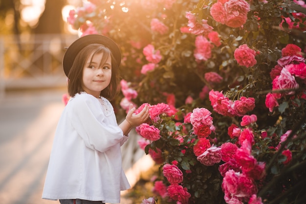 Una bambina carina ed elegante indossa abiti alla moda e cappello in posa su una rosa di fiori