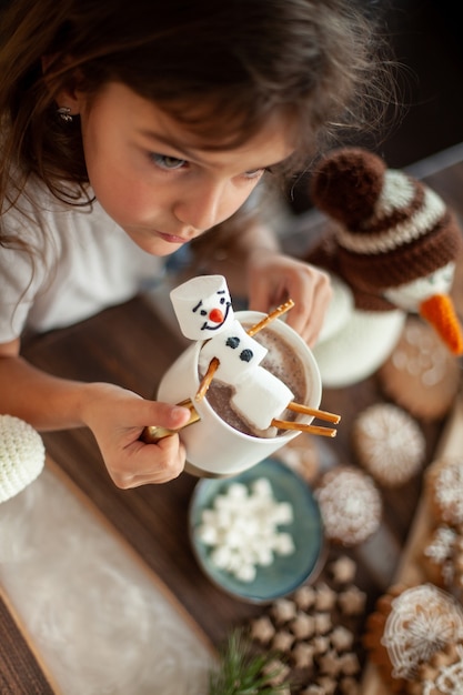 Una bambina carina gioca con pupazzi di neve a maglia e mangia pan di zenzero e beve cacao con marshmallow