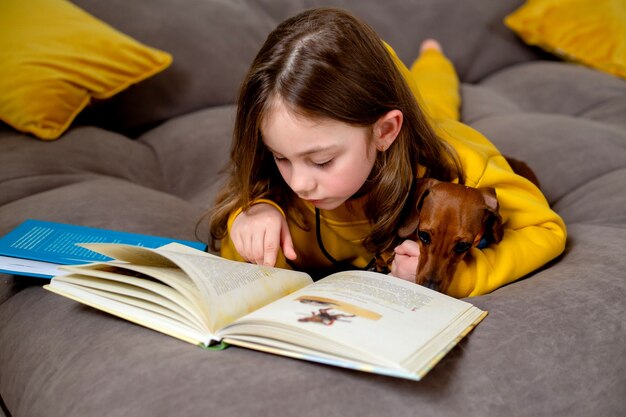 Una bambina carina in abiti gialli sta leggendo un libro sdraiato sul letto con un'educazione domestica del cane