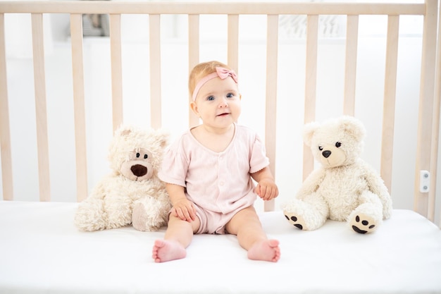 Una bambina carina in buona salute fino a un anno in un body rosa in tessuto naturale è seduta in una culla con orsacchiotti su lenzuola bianche in camera da letto guardando la telecamera il bambino è a casa