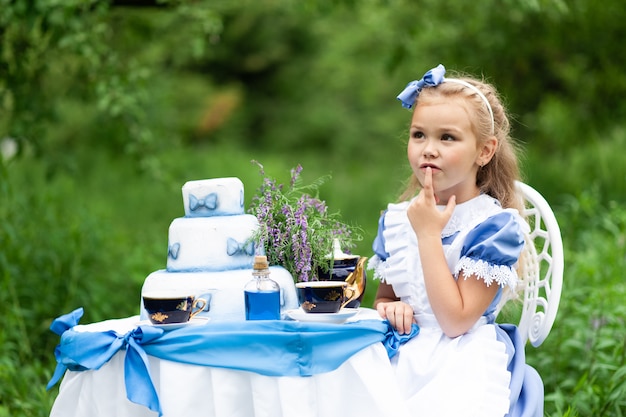 Una bambina carina in costume "Alice dal paese delle meraviglie" tiene un tea party al suo tavolo magico. Fotografato in natura.