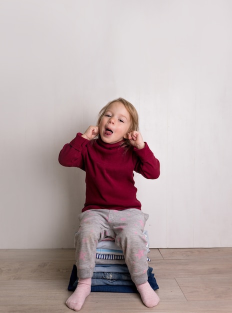 Una bambina carina in un maglione rosso si siede e sorride su una pila di vestiti piegati