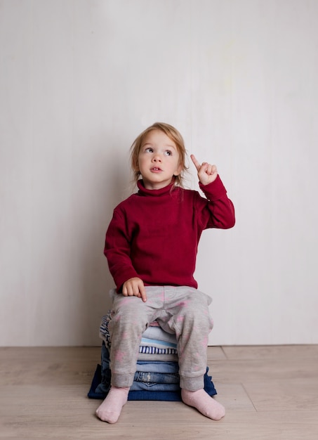 Una bambina carina in un maglione rosso si siede e sorride su una pila di vestiti piegati