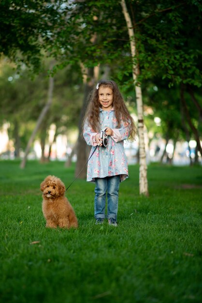 Una bambina carina in una passeggiata con il cane barboncino giocattolo. È primavera fuori. La ragazza è vestita con un mantello blu in un fiore e jeans. Ha lunghi capelli mossi. Infanzia. Passeggiare.