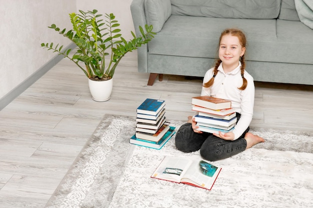 una bambina carina seduta a casa sul pavimento con una pila di libri nelle mani