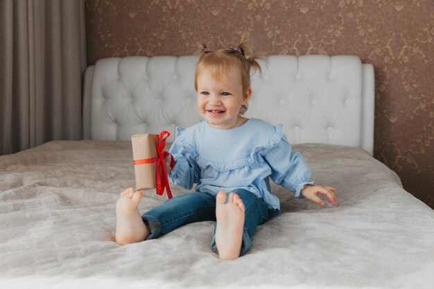 Una bambina carina si siede sul letto con una confezione regalo a casa, accetta gli auguri di compleanno.