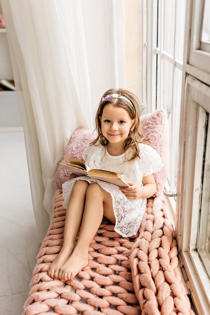 Una bambina carina sta leggendo un libro su un accogliente davanzale.