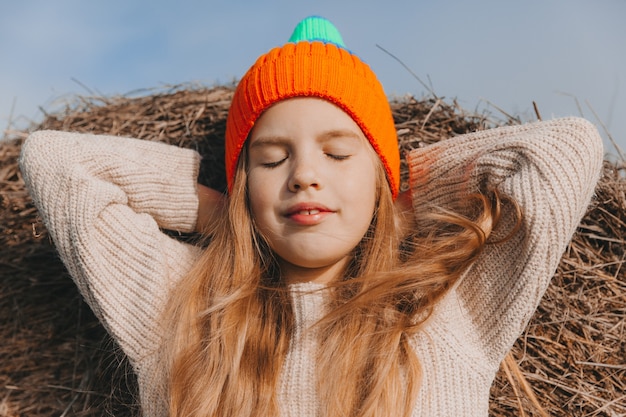 Una bambina con bei capelli biondi si siede su un campo vicino a un rotolo di fieno. ritratto di un bambino con un cappello a maglia.