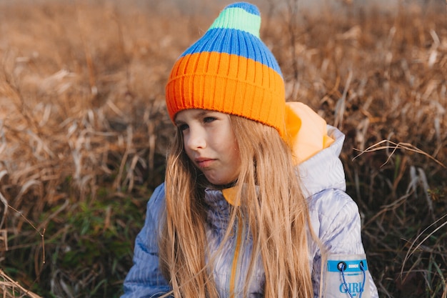 Una bambina con bei capelli biondi si siede su un campo vicino a un rotolo di fieno. ritratto di un bambino con un cappello a maglia.