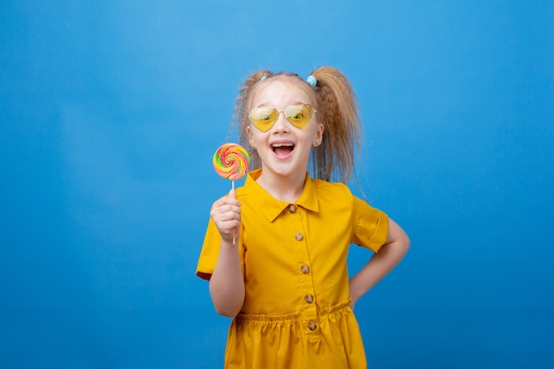 Una bambina con gli occhiali da sole tiene in mano un lecca-lecca su sfondo blu