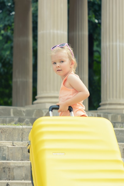 Una bambina con i capelli biondi solleva una grande valigia gialla sui gradini.