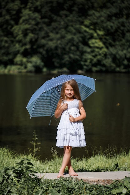 Una bambina con i capelli lunghi in abito bianco si trova sulla riva del fiume. Passeggiata in riva al lago in una giornata di sole estivo