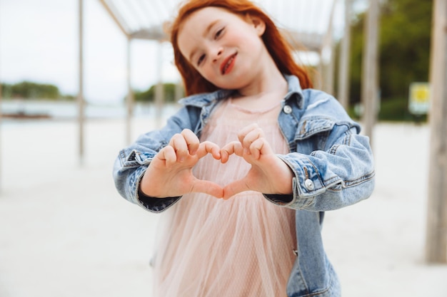 Una bambina con i capelli rossi sorride e mostra un segno del cuore con le mani un bambino sulla spiaggia