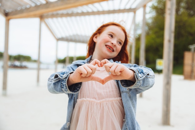 Una bambina con i capelli rossi sorride e mostra un segno del cuore con le mani un bambino sulla spiaggia
