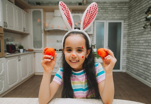 Una bambina con le orecchie da coniglio rosa tiene le uova di Pasqua sul viso vicino agli occhi in cucina