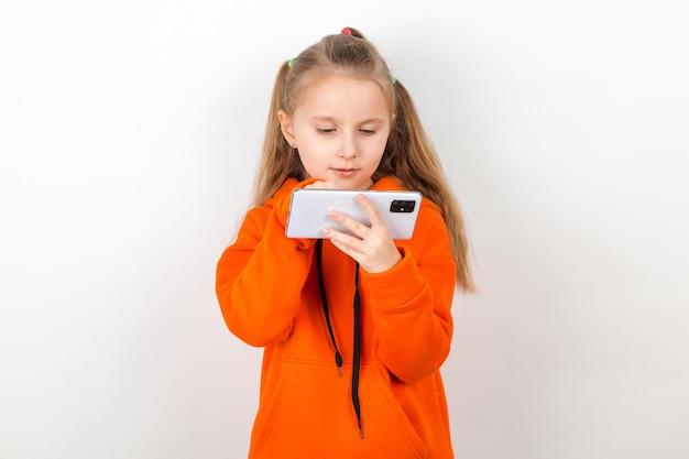 Una bambina con un abito arancione guarda il suo telefono Dipendenza di una generazione di bambini dai gadget Su uno sfondo bianco