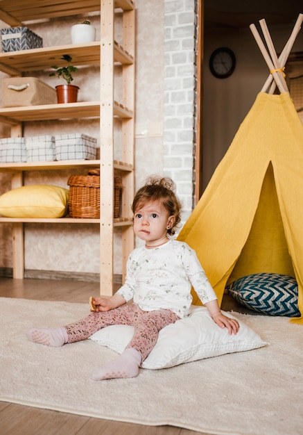 Una bambina con un biscotto in mano si siede sul pavimento con una faccia pensierosa. wigwam giallo