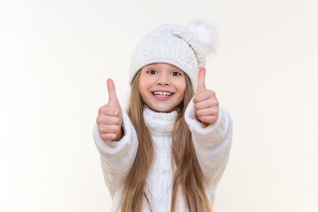 Una bambina con un cappello bianco e un maglione caldo mostra le sue dita in alto. Su uno sfondo bianco isolato.