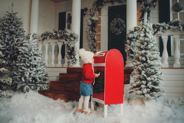 Una bambina con un cappello bianco lavorato a maglia esprime un desiderio e scrive una lettera a Babbo Natale e invia una cartolina per posta. Getta la busta nella cassetta della posta