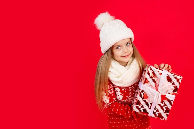 una bambina con un cappello d'inverno e un maglione con regali su uno sfondo rosso monocromo isolato gioisce e sorride il concetto di nuovo anno e spazio di Natale per il testo
