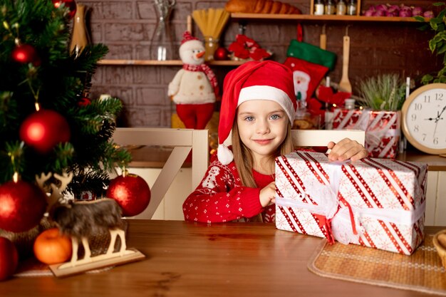 Una bambina con un cappello da Babbo Natale con doni in una cucina buia vicino a un albero di Natale con palline rosse esulta e sorride, il concetto di capodanno e Natale