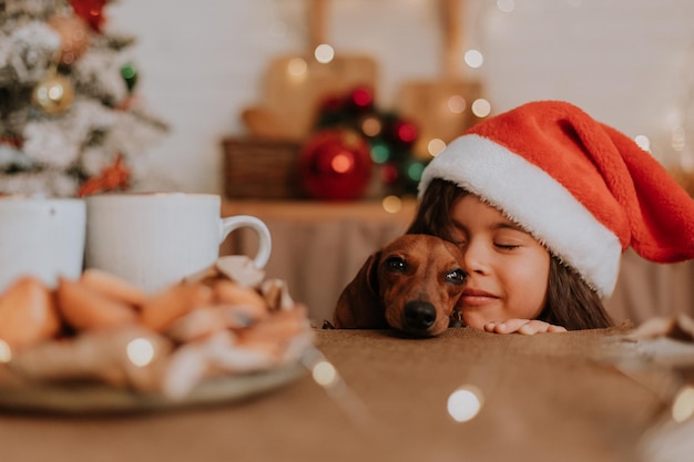 Una bambina con un cappello da Babbo Natale e un bassotto nano vogliono mangiare un piatto di dolci e una torta di Natale