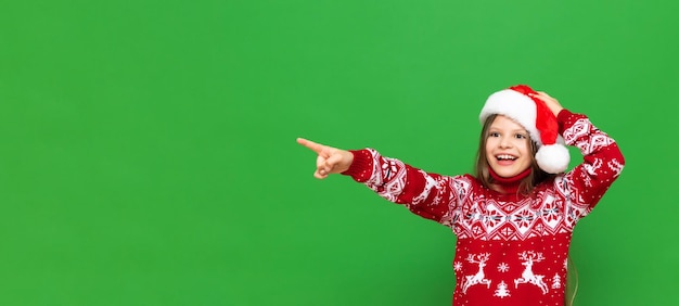 Una bambina con un cappello da Babbo Natale e un maglione festivo indica la tua pubblicità e sorride ampiamente su uno sfondo verde isolato Spazio di copia