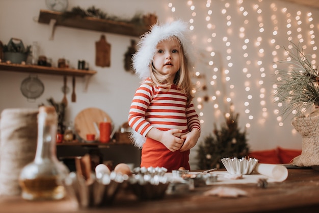 una bambina con un cappello da Babbo Natale e un pigiama a righe rosse cucina una torta di Natale in una bella cucina