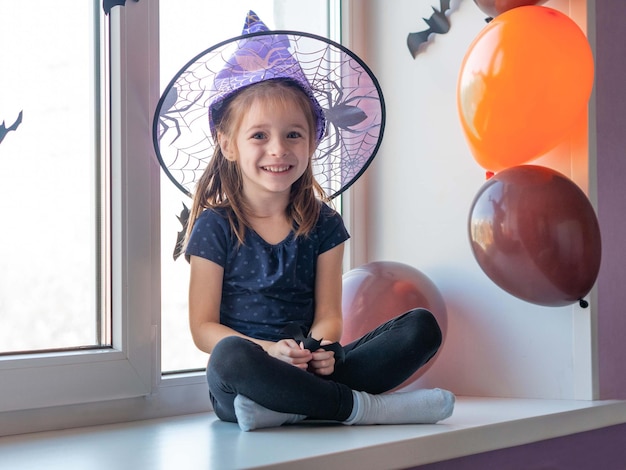 Una bambina con un cappello da strega è seduta sul davanzale felice Halloween