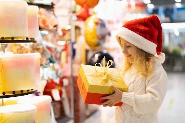 Una bambina con un cappello di natale con un regalo di capodanno in mano a una vetrina con giocattoli per bambini